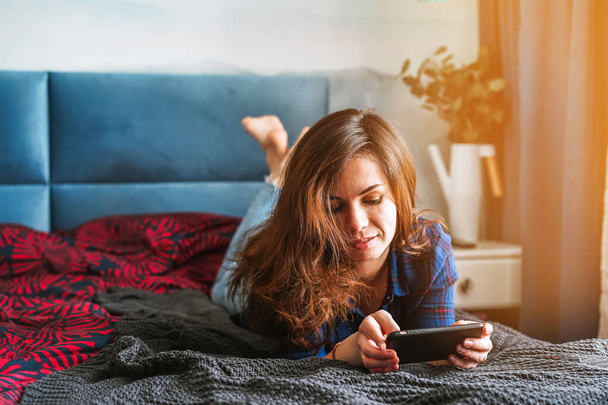 Μια μοντέρνα νεαρή γυναίκα με πουκάμισο και τζιν είναι ξαπλωμένη στο κρεβάτι στην κρεβατοκάμαρα και χρησιμοποιεί το smartphone της. - Φωτογραφία, εικόνα
