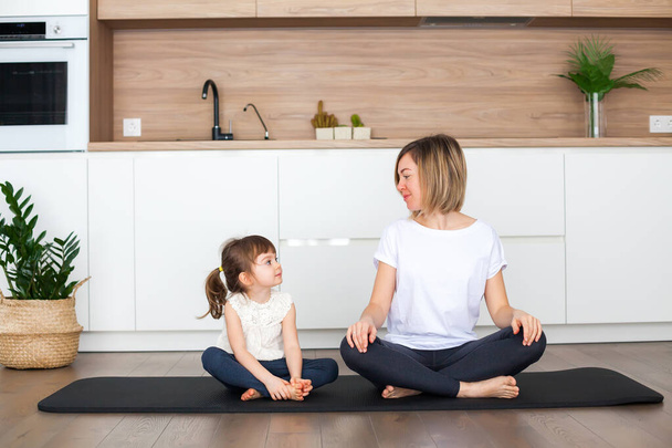 Woman and her cute little daughter are sitting in a lotus position while doing yoga together at home - Fotoğraf, Görsel