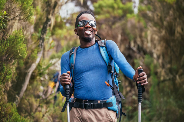 Portrait of a cheerfully smiling African-American Ethnicity young man in sunglasses. He having a walk with a backpack using trekking poles in the forest. Active people and traveling concept.  - Φωτογραφία, εικόνα