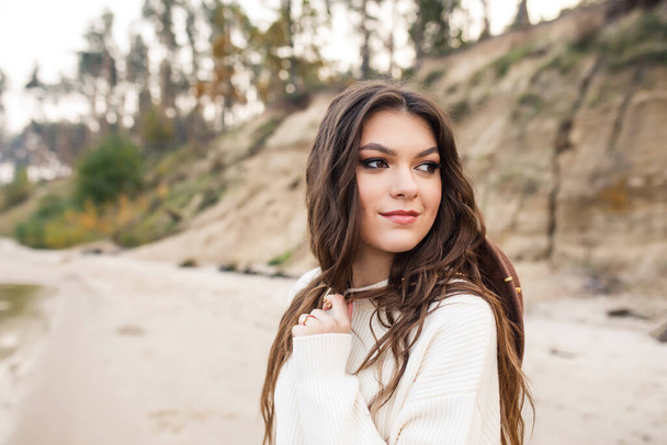 young beautiful girl in a white sweater, blue jeans and brown hat are walking on the coast near the lake - Foto, imagen