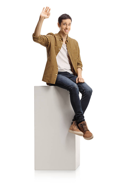 Shy young man sitting on a white column and waving isolated on white background - Foto, afbeelding