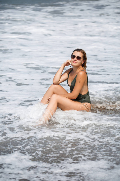 Woman in sunglasses in a green swimsuit stands on the sand by the ocean and has a rest - Foto, Imagem