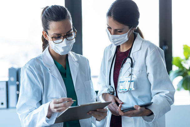 Prise de vue de deux jeunes femmes médecin portant un masque facial hygiénique parlant avec son collègue tout en travaillant tablette numérique à l'hôpital. - Photo, image