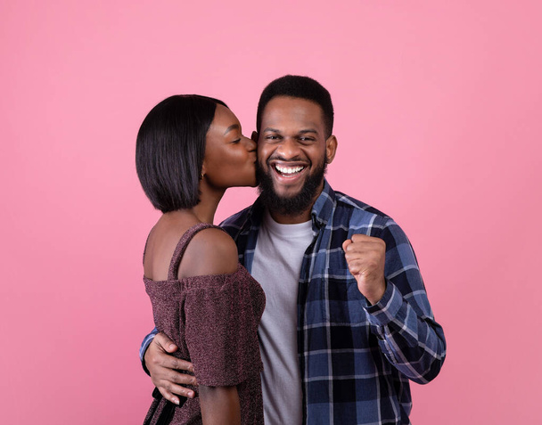Excited black guy making YES gesture, young girlfriend kissing his cheek on pink studio background. Valentines Day - Foto, Bild