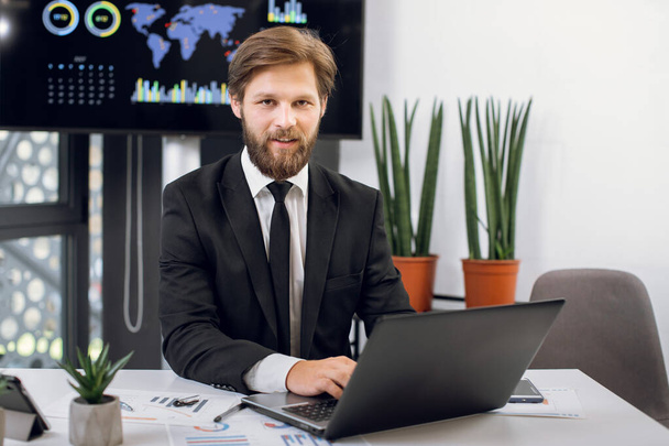 Νέος σκόπιμα χαμογελαστός γενειοφόρος αφεντικό ή διευθύνων σύμβουλος σε μαύρο κοστούμι, κάθεται στο χώρο εργασίας του στο σύγχρονο γραφείο και εργάζονται σε φορητό υπολογιστή, κοιτάζοντας την κάμερα - Φωτογραφία, εικόνα