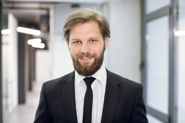 Happy young positive male office worker manager, wearing black suit and tie, smiling at camera, standing on the background of modern light office corridor. Close up face portrait - Foto, Bild