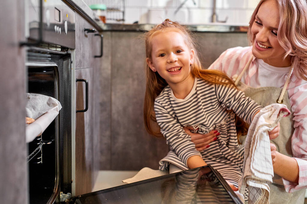 pretty daughter and mother taking out tray of baked cookies from oven in kitchen - Zdjęcie, obraz