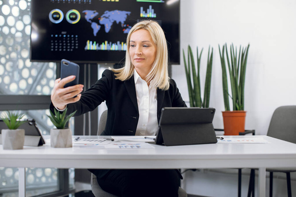 Successful charming blond haired mature lady economist in smart outfit, making photo on her phone camera, while sitting at the desk in modern office with wall plasma screen on the background - Фото, зображення