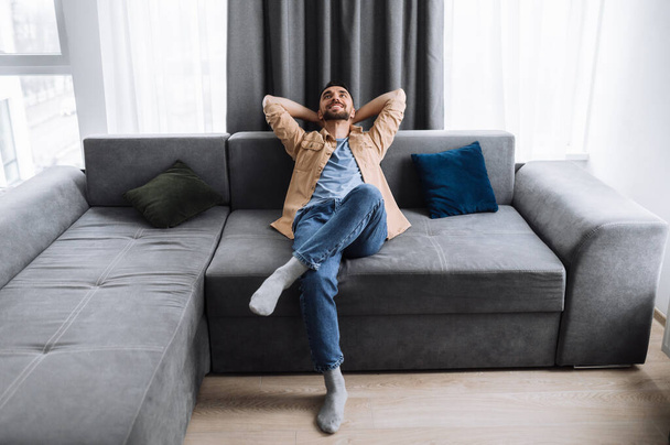 Zufriedener gutaussehender kaukasischer Mann sitzt auf der Couch im Wohnzimmer, denkt über Pläne für das Wochenende oder den Urlaub nach und lächelt. Junge Freiberufler ruhen sich von Online-Arbeit aus - Foto, Bild