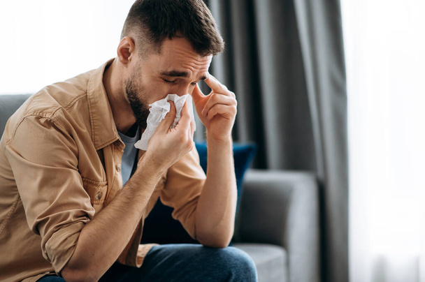 Unhealthy young man. Caucasian guy is sick with flu, he sneezes and blows his nose in a napkin while sitting on the couch at home - Фото, изображение