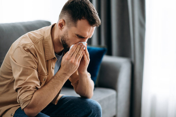 Sick caucasian guy sits on couch feel unhealthy, he suffering from rhinitis snuffles, having respiratory infection, get flue, sitting at home at quarantine - Фото, зображення