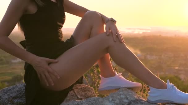 Close-up van jonge elegante vrouwelijke slanke benen in zwart korte jurk zitten op een rots ontspannen buiten in de zomer avond. Modieuze vrouw bij warme zonsondergang in de natuur. - Video