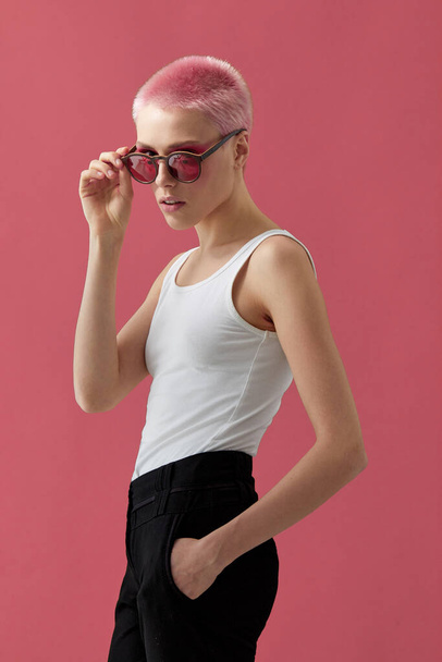 Hipster κορίτσι με κοντά ροζ μαλλιά, φορώντας λευκό μπλουζάκι και μαύρο παντελόνι και γυαλιά ηλίου με ξύλινο σκελετό και κόκκινους φακούς, πάνω από ροζ φόντο. Κάθετη προβολή. - Φωτογραφία, εικόνα