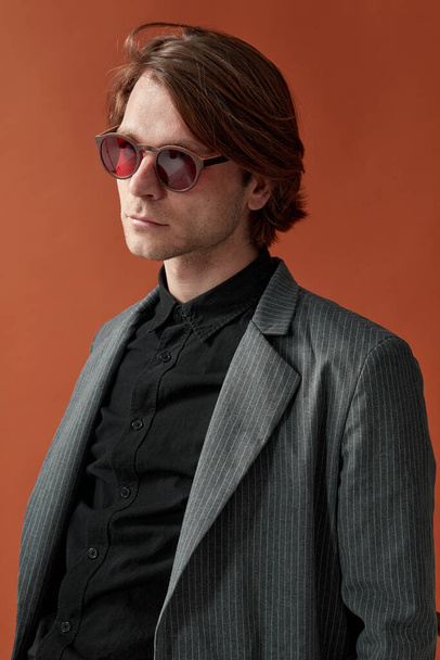 Atractivo modelo masculino elegante en gafas rojas, de pie con una postura firme sobre fondo de estudio marrón. Vista vertical. - Foto, imagen