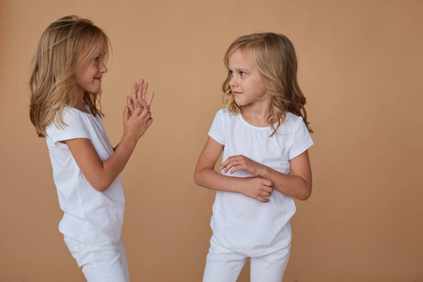 Front portrait of adorable little twin sisters with wavy blond hair, talk each other, wears white clothes, over beige background. Horizontal view. - Photo, Image
