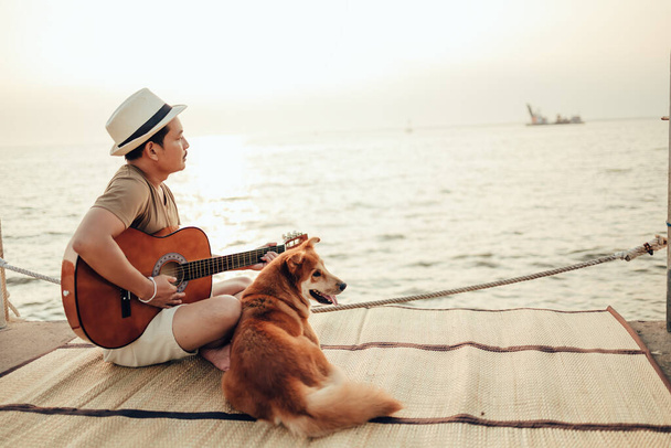 A man wear straw hat and playing guitar music song near the sea sunset with a dog pet - Foto, immagini
