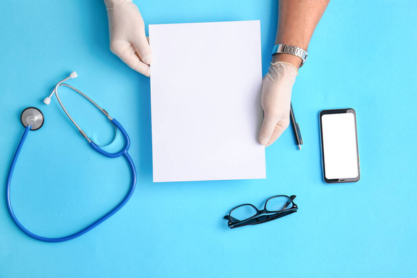 Mužské ruce v lékařských rukavicích s prázdným papírovým listem a stetoskopem na barevném pozadí - Fotografie, Obrázek