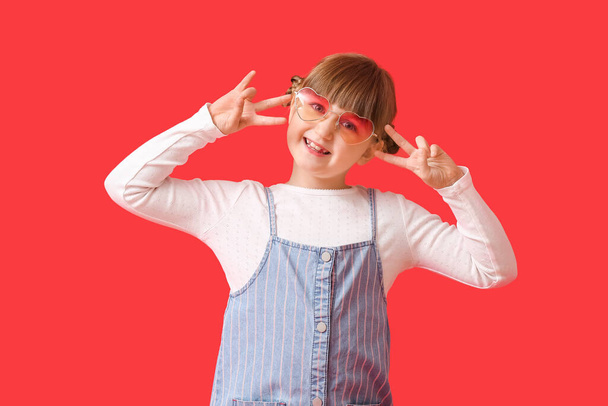 Linda niña con gafas de sol elegantes que muestran el gesto de victoria en el fondo de color - Foto, Imagen