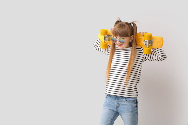 Χαριτωμένο κοριτσάκι με κομψά γυαλιά ηλίου και skateboard στο φως φόντο - Φωτογραφία, εικόνα