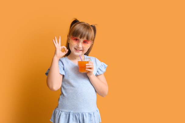 Милая маленькая девочка в стильных солнцезащитных очках и стакане сока показывает ОК жеста на цветном фоне - Фото, изображение
