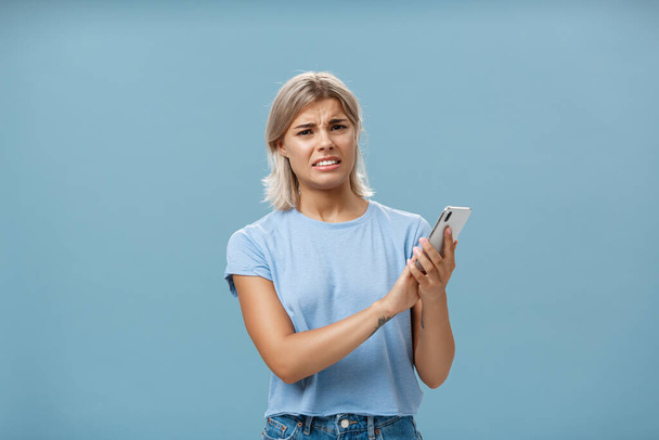 Woman cannot stand when mom shouts during phone call. Intense displeased young european blonde with tanned skin clenching teeth and frowning covering microphone of smartphone over blue wall - Photo, image