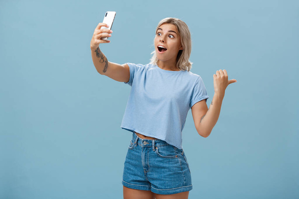 Girl recording video showing followers awesome scene attending cool event holding smartphone looking impressed and surprised at device screen pointing backwards indicating right over blue wall - Foto, Imagen