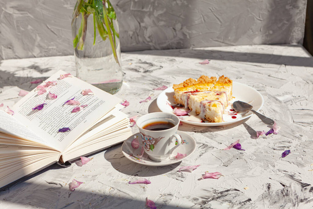 Ένα ανοιχτό βιβλίο βρίσκεται δίπλα σε μια πίτα και ένα φλιτζάνι καφέ σε ένα γκρι φόντο σκορπισμένα με πέταλα λουλουδιών. Υπέροχο πρωινό. - Φωτογραφία, εικόνα