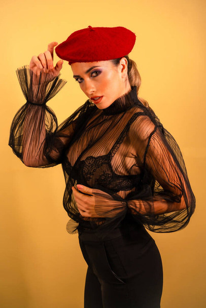 A sexy young female in a red barret hat and a black transparent shirt posing at camera - Zdjęcie, obraz