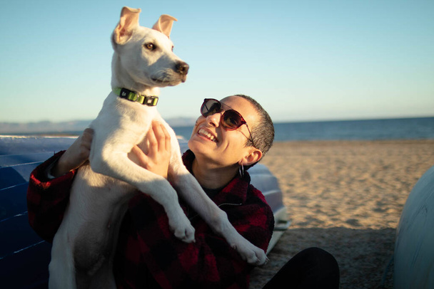 Un plano superficial de una hembra con gafas de sol jugando con su perro blanco en la playa - Foto, imagen