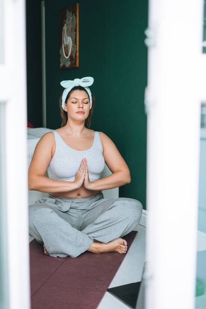 Mooie brunette jonge vrouw plus size lichaam positief in comfortabele sport praktijk yoga online thuis - Foto, afbeelding