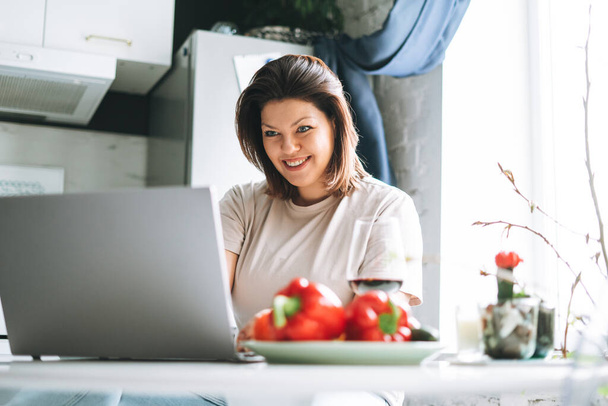 Όμορφη χαμογελαστή μελαχρινή νεαρή γυναίκα συν το μέγεθος του σώματος θετική χρησιμοποιώντας φορητό υπολογιστή στην κουζίνα στο σπίτι - Φωτογραφία, εικόνα