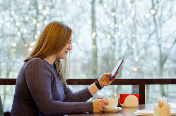 junge Frau im Café trinkt Kaffee und nutzt Smartphone und Wifi, verbindet sich mit Freunden, lädt App oder Anwendung aus dem Internet herunter, verbringt Zeit mit modernem Smartphone, liest Nachrichten, chattet - Foto, Bild