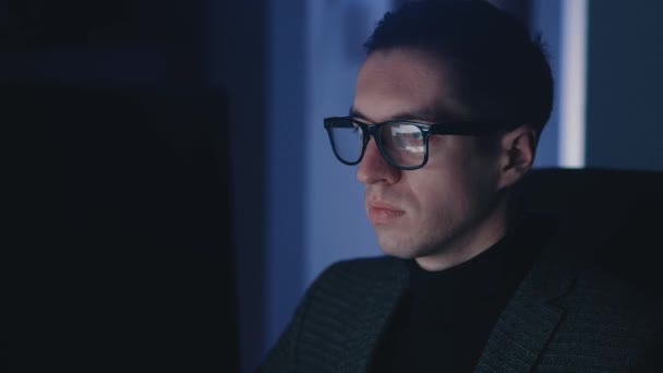 Egy fiatal üzletember, szemüveges szabadúszó, aki késő este dolgozik a laptopnál. Portré a sikeres férfi vállalkozó szörfözés internet éjjel, tükröződik monitor az arcán - Felvétel, videó