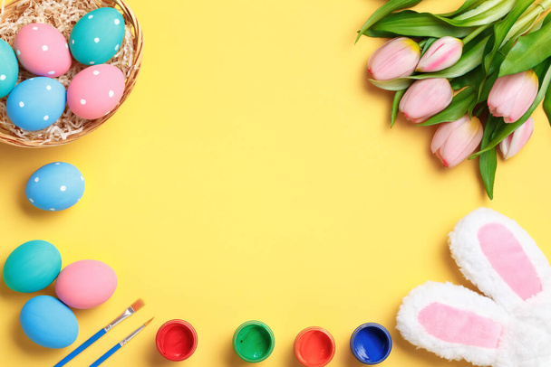 Happy easter. Multi-colored eggs in a basket, rabbit ears, tulips and paints on a yellow background. Flat lay greeting card. Copy space on medical protective mask. - Фото, зображення