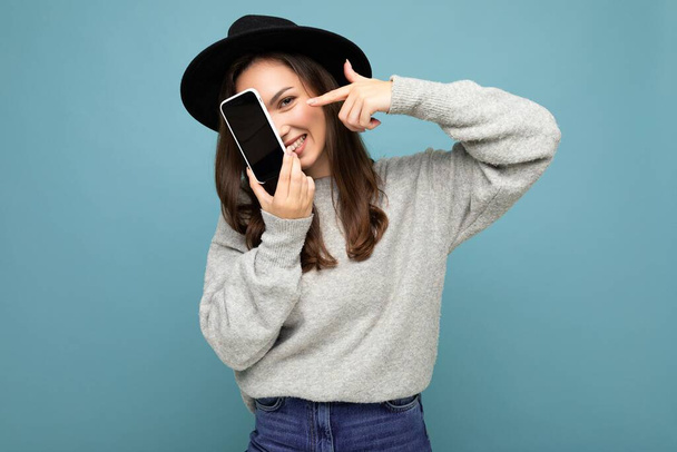 Pretty young smiling woman wearing black hat and grey sweater holding phone looking at camera isolated on background - Photo, Image