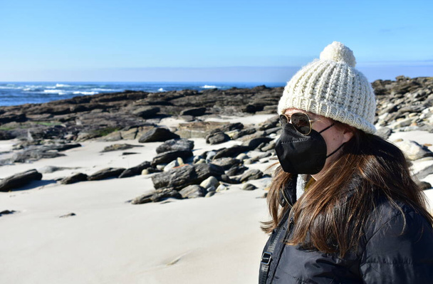 Donna con maschera nera Covid-19 su una spiaggia in inverno con cappotto nero, occhiali da sole e cappello di lana bianca. Maschera KN95 o N95 o FFP2. - Foto, immagini