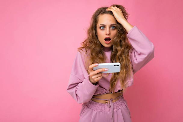 Zdjęcie atrakcyjnej szalonej zaskoczonej młodej kobiety ubranej w stylowe ubrania stojącej w izolacji nad tłem z kopią przestrzeni trzymającej i korzystającej z telefonu komórkowego patrzącego w kamerę - Zdjęcie, obraz