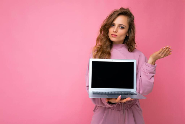 Close-up portrait of beautiful young dark blond curly hair woman holding computer laptop with empty copy space looking at camera and thinking about something having doubts posing hand isolated on pinl - Fotó, kép