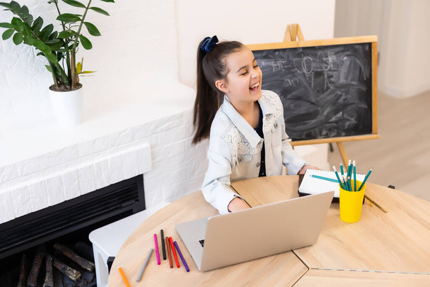 Smart little Caucasian girl child sit at table study at laptop make notes write in notebook, small schoolgirl handwrite prepare homework assignment at home, distant education concept - Photo, Image