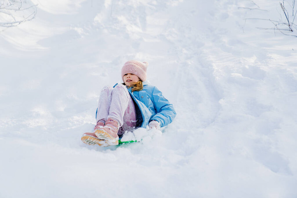 Een tiener meisje in een blauwe jas probeert te bewegen op een slee van een sneeuw glijbaan - Foto, afbeelding