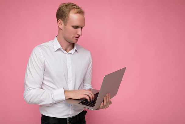 Zijprofiel foto shot van knappe zelfverzekerde blonde man met computer laptop typen op toetsenbord dragen witte shirt kijken naar monitor geïsoleerd over roze achtergrond - Foto, afbeelding