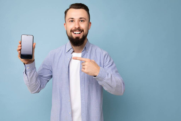 Photo of handsome smiling adult male person good looking wearing casual outfit standing isolated on background with copy space holding smartphone showing phone in hand with empty screen display for - Foto, Imagen