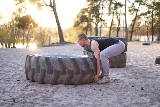Strong man training workout lifting large tire outdoor DIY gym. Sportive young adult caucasian guy flip big wheel on nature. Handmade equipment sport exercising helthy lifestyle concept - Φωτογραφία, εικόνα