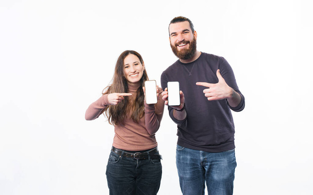Lächelndes junges Paar steht vor weißem Hintergrund und zeigt auf Smartphone-Bildschirme - Foto, Bild