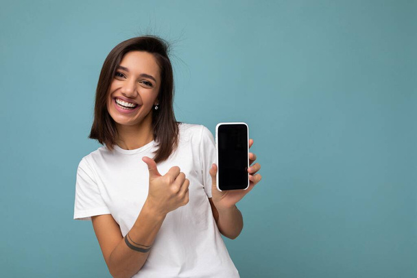 Attractive smiling young brunet woman good looking wearing white t-shirt standing isolated on blue background with copy space holding smartphone showing phone in hand with empty screen screen for - Foto, Imagen