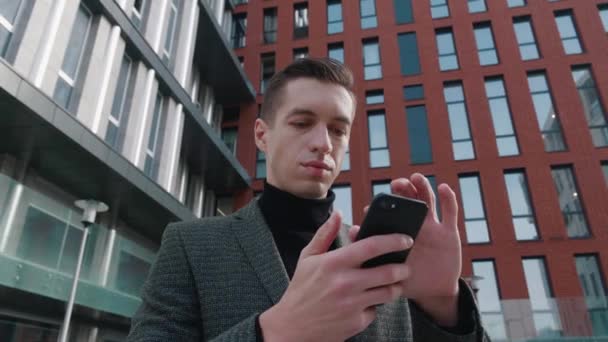 Portret van lachende man zakenman staan in het centrum van de stad straat en met behulp van smartphone. Jonge ondernemer in pak met behulp van mobiele telefoon buiten op moderne gebouw achtergrond. - Video