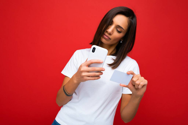 Όμορφη γυναίκα φορώντας καθημερινά κομψά ρούχα που απομονώνονται σε φόντο τοίχο κρατώντας και χρησιμοποιώντας τηλέφωνο και πιστωτική κάρτα κάνοντας την πληρωμή κοιτάζοντας οθόνη smartphone - Φωτογραφία, εικόνα