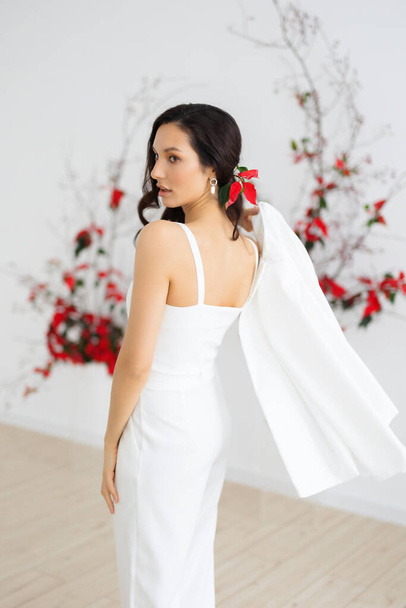 A young caucasian brunette woman in a stylish and elegant pantsuit holding a jacket. Bride posing against the background of an unusual composition of fresh flowers poinsetia for an chamber wedding ceremony - Фото, зображення