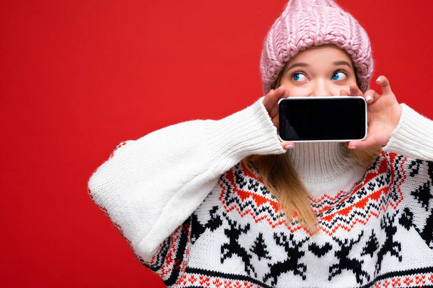 Photo of attractive shocked young blonde woman wearing warm knitted hat and winter warm sweater standing isolated over red background showing smartphone with empty screen for cutout looking to the - Фото, зображення