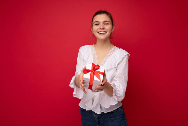 Beautiful happy young brunette woman isolated over colourful background wall wearing stylish casual clothes holding gift box and looking at camera - Foto, immagini